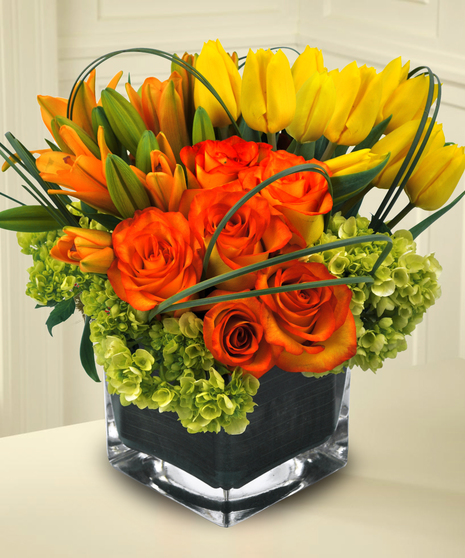 Orange lilies, yellow tulips, green hydrangea and orange roses in a leaf-lined cube vase.