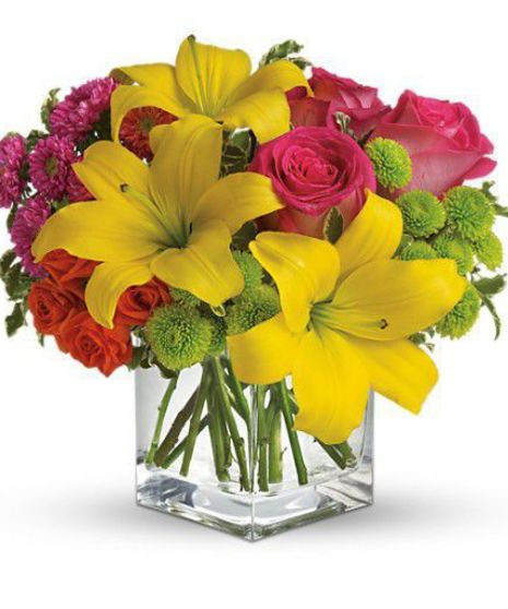 Hot pink roses, orange spray roses, yellow asiatic lilies, hot pink matsumoto asters and green button spray chrysanthemums in a cube vase.